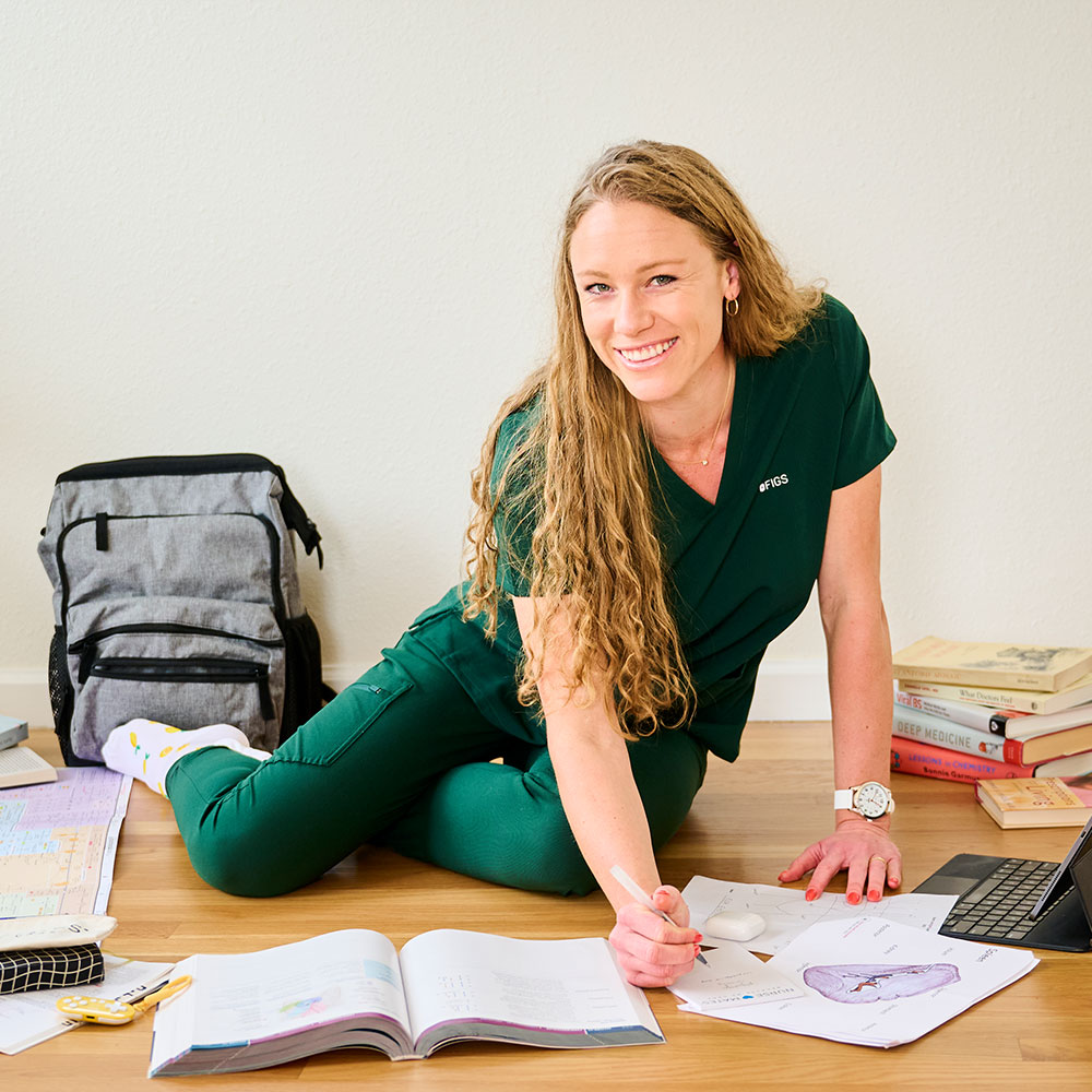 Ella is sitting on the floor surrounded by textbooks and study materials, wearing green scrubs, Lemon Squeezy Compression Socks, and has the Ultimate Nursing Bag in Grey Linen behind her.